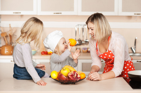Little cute girl with sister smiling and showing orange to their mother while helping to cook salad. Kitchen interior. Concept for young kitchen handsの写真素材