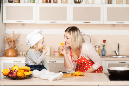 Little cute girl with her mother eating orange while cooking. Kitchen interior. Concept for young kitchen handsの写真素材