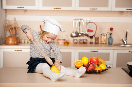 Little cute girl with chef hat holding big wooden spoon while cooking. Kitchen interior. Concept for young kitchen handsの写真素材