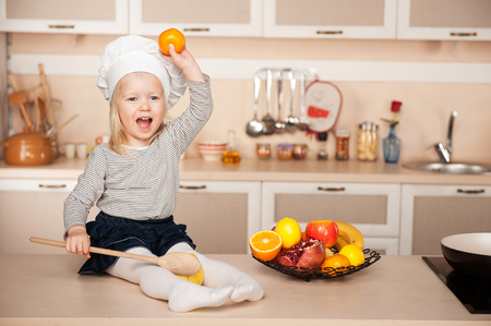 Little cute girl with chef hat and big wooden spoon holding apple. Kitchen interior. Concept for young kitchen handsの写真素材