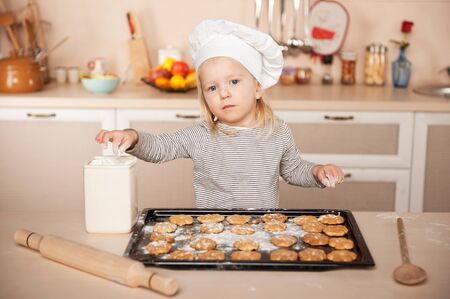 Little cute girl with chef hat looking at camera and preparing cookies. Kitchen interior. Concept for young kitchen handsの写真素材