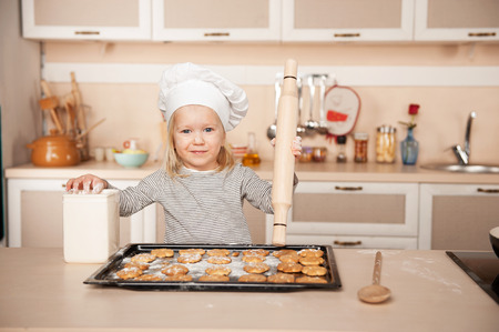 Little cute girl with chef hat and rolling pin looking at camera and preparing cookies. Kitchen interior. Concept for young kitchen handsの写真素材