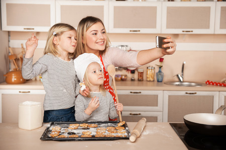 Little cute girls and their mother making photo while preparing cookies. Kitchen interior. Concept for young kitchen handsの写真素材