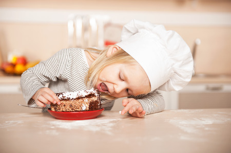 Little cute girl with chef hat eating cake. Kitchen interior. Concept for young kitchen handsの写真素材