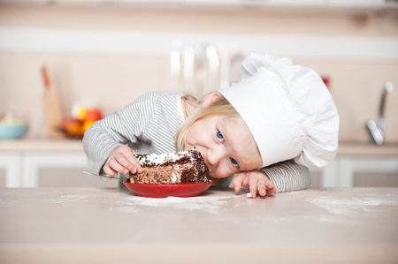 Little funny girl with chef hat eating cake and looking at camera. Kitchen interior. Concept for young kitchen handsの写真素材