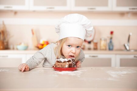Little funny girl with chef hat eating cake. Kitchen interior. Concept for young kitchen handsの写真素材