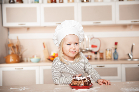 Little funny girl with chef hat and mouth full of cake. Kitchen interior. Concept for young kitchen handsの写真素材