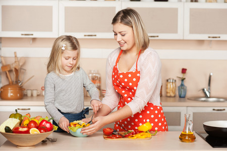 Beautiful smiling young mother and her little daughter cooking salad. Kitchen interior. Concept for young kitchen handsの写真素材