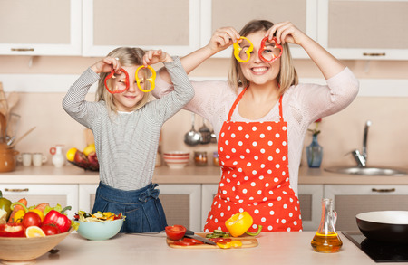 Beautiful smiling young mother and her little daughter having fun while cooking salad. Kitchen interior. Concept for young kitchen handsの写真素材