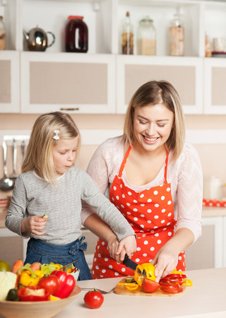 Beautiful smiling young mother helping her little daughter to chop paprika while cooking salad. Kitchen interior. Concept for young kitchen handsの写真素材