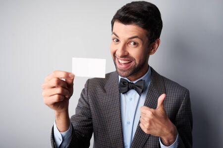 Portrait of positive casual young man wearing jacket and bow tie. Man smiling, showing thumb up and holding blank visiting cardの写真素材