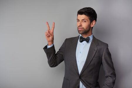 Portrait of elegant casual young man wearing tweed jacket and bow tie. Man showing victory sign and looking at cameraの写真素材