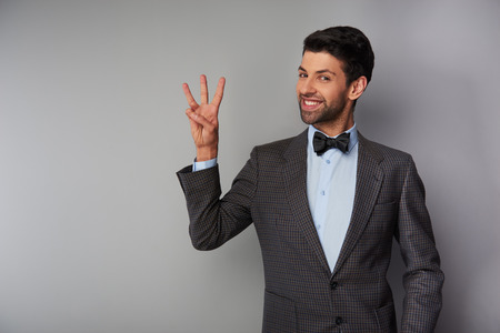 Portrait of positive casual young man wearing tweed jacket and bow tie. Man smiling, showing three fingers and looking at cameraの写真素材