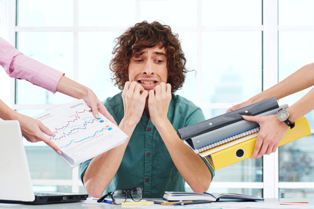 Scared young businessman looking at camera. Hands with folders around him. Office interior with window. Concept for overworkingの写真素材