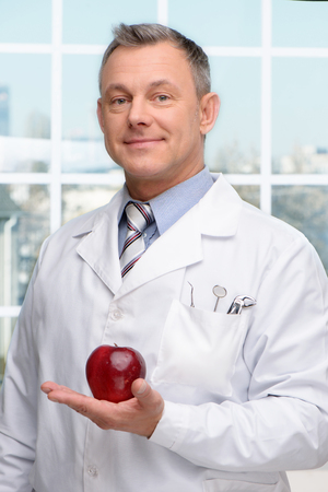 Photo of dentist holding red apple and looking at camera. Concept for teeth hygieneの写真素材