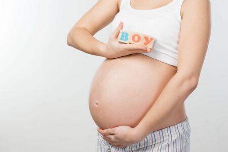 Close up photo of young pregnant woman belly. Woman holding wooden cubes with word "boy" painted on it. Isolated on white background. Concept for pregnancyの写真素材