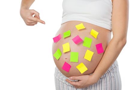 Close up photo of young pregnant woman. Woman standing isolated on white background and pointing at colorful paper stickers on her belly. Concept for pregnancyの写真素材