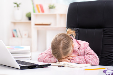 Funny picture of little cute girl playing role of business woman. Girl wearing pink suit. Girl sitting at table on large office chair and sleeping near laptop. Office interior as a backgroundの写真素材