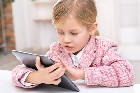 Portrait of little cute girl wearing pink suit. Girl sitting at table and using tablet computer. Room interior as a backgroundの写真素材