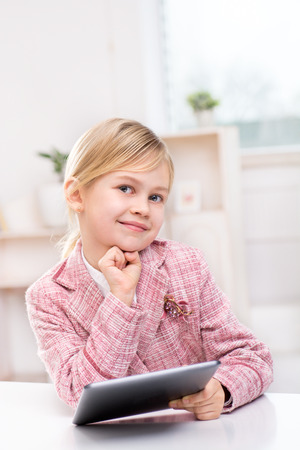 Portrait of little cute girl wearing pink suit. Girl sitting at table, smiling and holding tablet computer. Room interior as a backgroundの写真素材