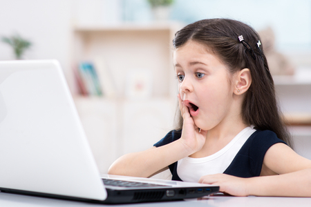 Nice picture of little dark-haired girl. Girl sitting at table and looking at laptop in amazement. Room interior as a backgroundの写真素材