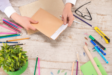 Creative top view photo of man hands holding envelope with letter. Multi-colored pencils and markers, straight edge tool, paper clips and knife, glasses, green plant are on the light-colored woodblocksの写真素材