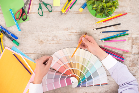 Creative top view photo of man hands pointing at color palette. Multi-colored pencils and markers, paper knife, green plant, color paper, scissors are on the light-colored woodblocksの写真素材