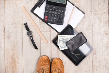 Creative top view photo of vintage brown shoes near paper, watches, purse with money, pencil, calculator, straight edge tool and tablet computer on the light-colored woodblocksの写真素材