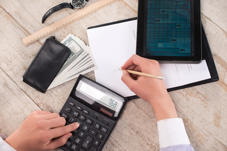 Creative top view photo of young bisinessman counting with calculator and writing numbers on paper. Man sitting near watches, purse with money, straight edge tool and tablet computer on the light-colored woodblocksの写真素材