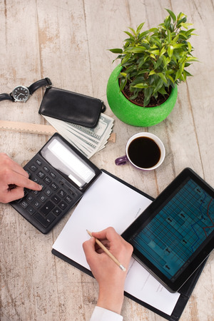 Creative vertical top view photo of young bisinessman counting with calculator and writing numbers on paper. Man sitting near watches, purse with money, straight edge tool, green plant, cup of coffee and tablet computer on the light-colored woodblocksの写真素材