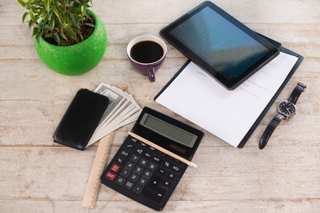 Creative top view photo of paper, watches, purse with money, pencil, calculator, straight edge tool, green plant, cup of coffee and tablet computer on the light-colored woodblocksの写真素材