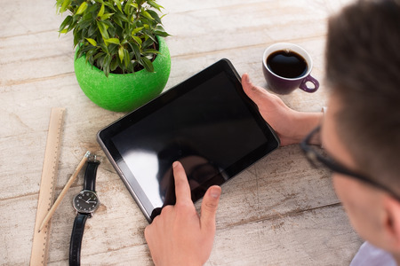 Creative top view photo of young bisinessman working with tablet computer. Man sitting near watches, straight edge tool, green plant and cup of coffee on the light-colored woodblocksの写真素材