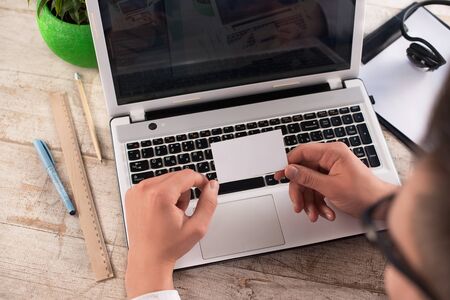 Creative top view photo of young bisinessman holding blank white visit card near laptop, head-phones, straight edge tool, green plant and pancils on the light-colored woodblocksの写真素材