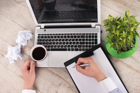 Creative top view photo of man hand with laptop, cup of coffee, green plant and crumpled paper on the light-colored woodblocksの写真素材