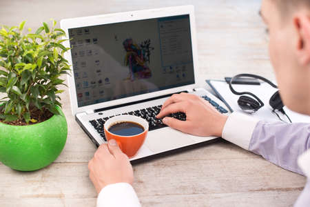 Creative top view photo of young businessman using laptop and holding cup of coffee. Man sitting near green plant on the light-colored woodblocksの写真素材