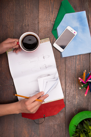 Creative vertical top view photo of woman hand with cup of coffee, paper, green plant, colored pencils, tissue pieces and phone on the dark-colored woodblocksの写真素材