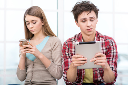 Handsome boy and beautiful girl using phone and tablet computer. They sitting in light room with large window. Concept for teenagersの写真素材