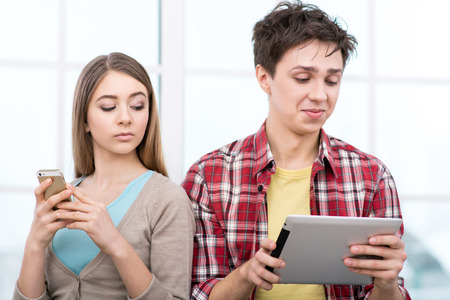 Handsome boy and beautiful girl using phone and tablet computer. They sitting in light room with large window. Girl spying for boy. Concept for teenagersの写真素材