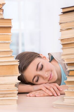 Nice photo of beautiful sleeping girl on desk. Girl is in light room with large window near stacks of books. Concept for teenagersの写真素材