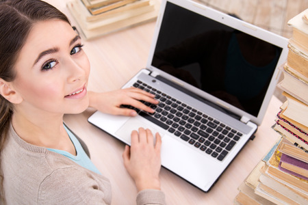 Top view photo of beautiful girl. She working with laptop, looking at camera and sitting near stacks of books. Concept for teenagersの写真素材