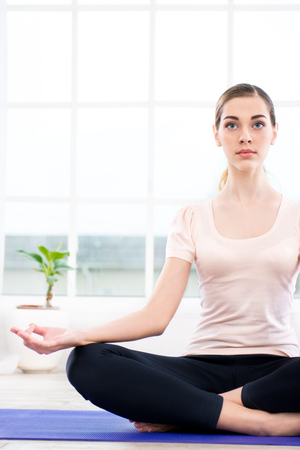 Nice photo of young woman practicing yoga. Woman meditating while doing lotus pose. White interior with large windowの写真素材