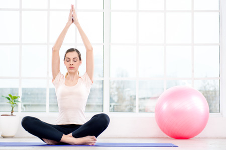 Nice photo of young woman practicing yoga. Woman meditating with her eyes closed while doing lotus pose. White interior with large window and pink fitness ballの写真素材
