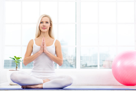 Nice photo of beautiful woman practicing yoga. Woman meditating while doing lotus pose. White interior with large window and pink fitness ballの写真素材