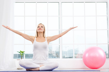 Nice photo of beautiful woman practicing yoga. Woman meditating with her hands up while doing lotus pose. White interior with large window and pink fitness ballの写真素材