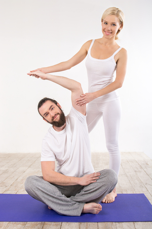 Nice photo of beautiful sporty woman and man. Woman helping man with exersises on violet fitness mat and smiling. White interiorの写真素材