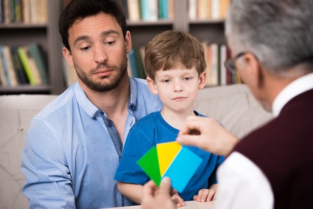 Little boy and his father talking with psychologist. Psychologist making test with boy. There are many books in psychologist officeの写真素材