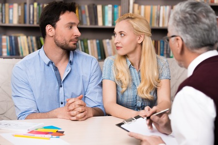 Young couple talking with psychologist. Psychologist taking notes. Man and woman looking at each other. There are many books in psychologist officeの写真素材