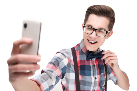 Young nerd in glasses and checkered shirt with smartphone making selfie isolated on white backgroundの写真素材