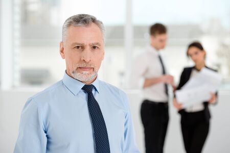Aged businessman looking at camera. Business people communicating in the backgroundの写真素材