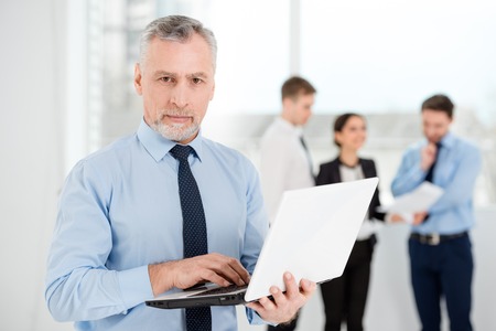 Aged businessman using white laptop and looking at camera. Business people communicating in the backgroundの写真素材